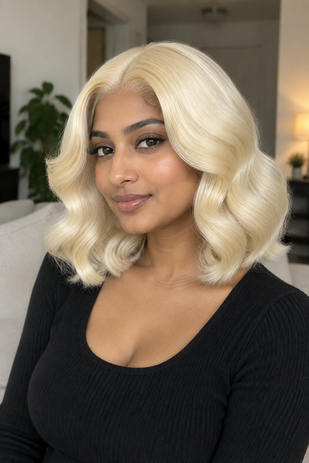 A woman wearing a shoulder-length, wavy blonde wig with a natural-looking part, styled with soft curls, paired with a black ribbed top in an indoor setting.