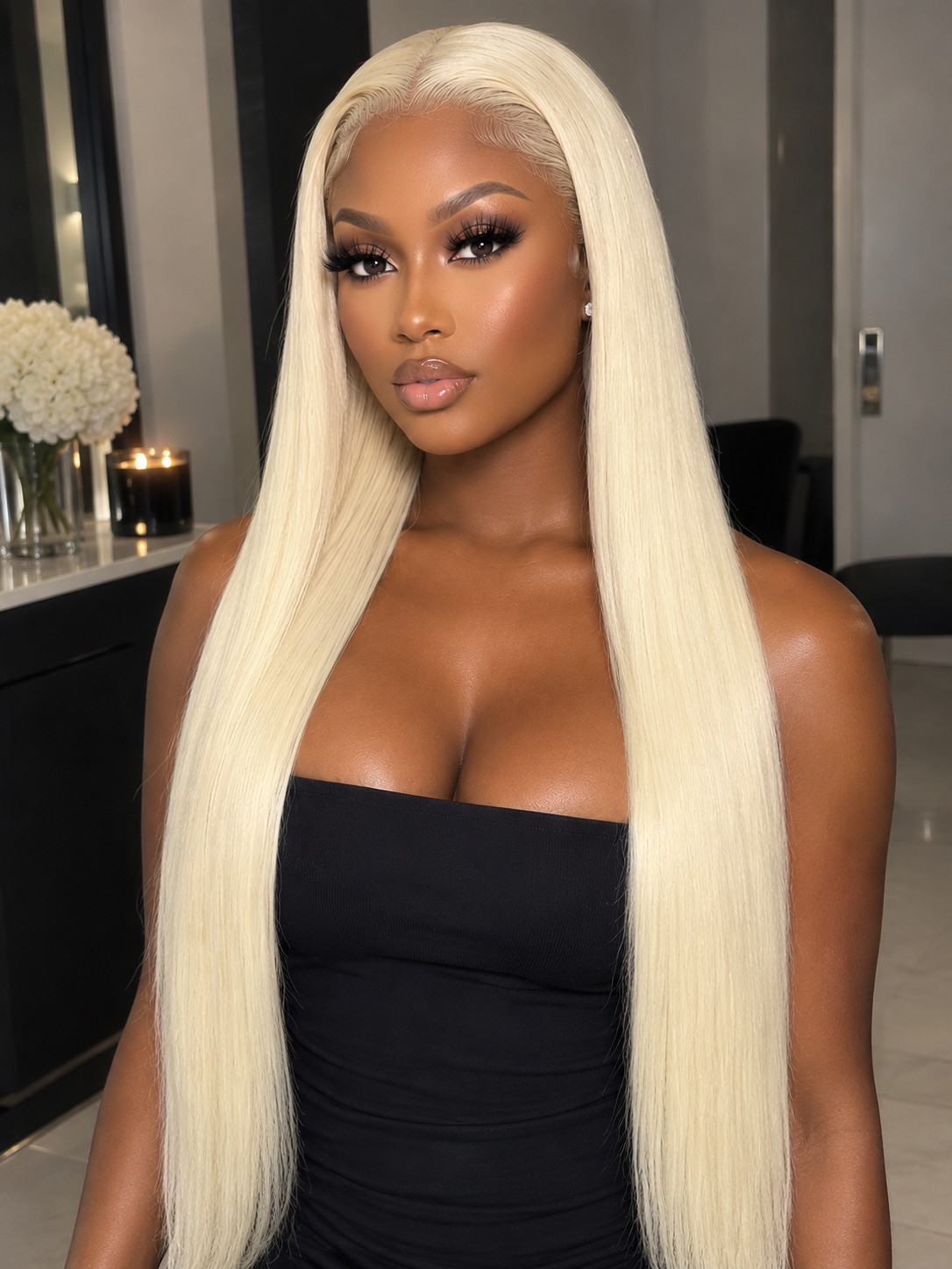 A woman with long, straight platinum blonde hair wearing a black strapless top, photographed indoors with a blurred modern room background.