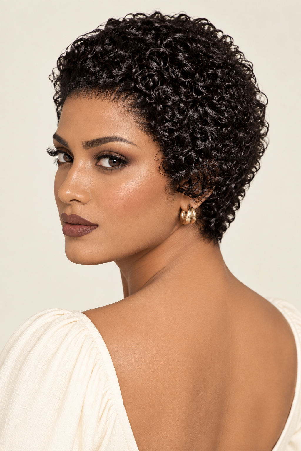 A woman with short, curly black hair shown from the back and side, wearing a cream off-the-shoulder top and gold hoop earrings against a light beige background.