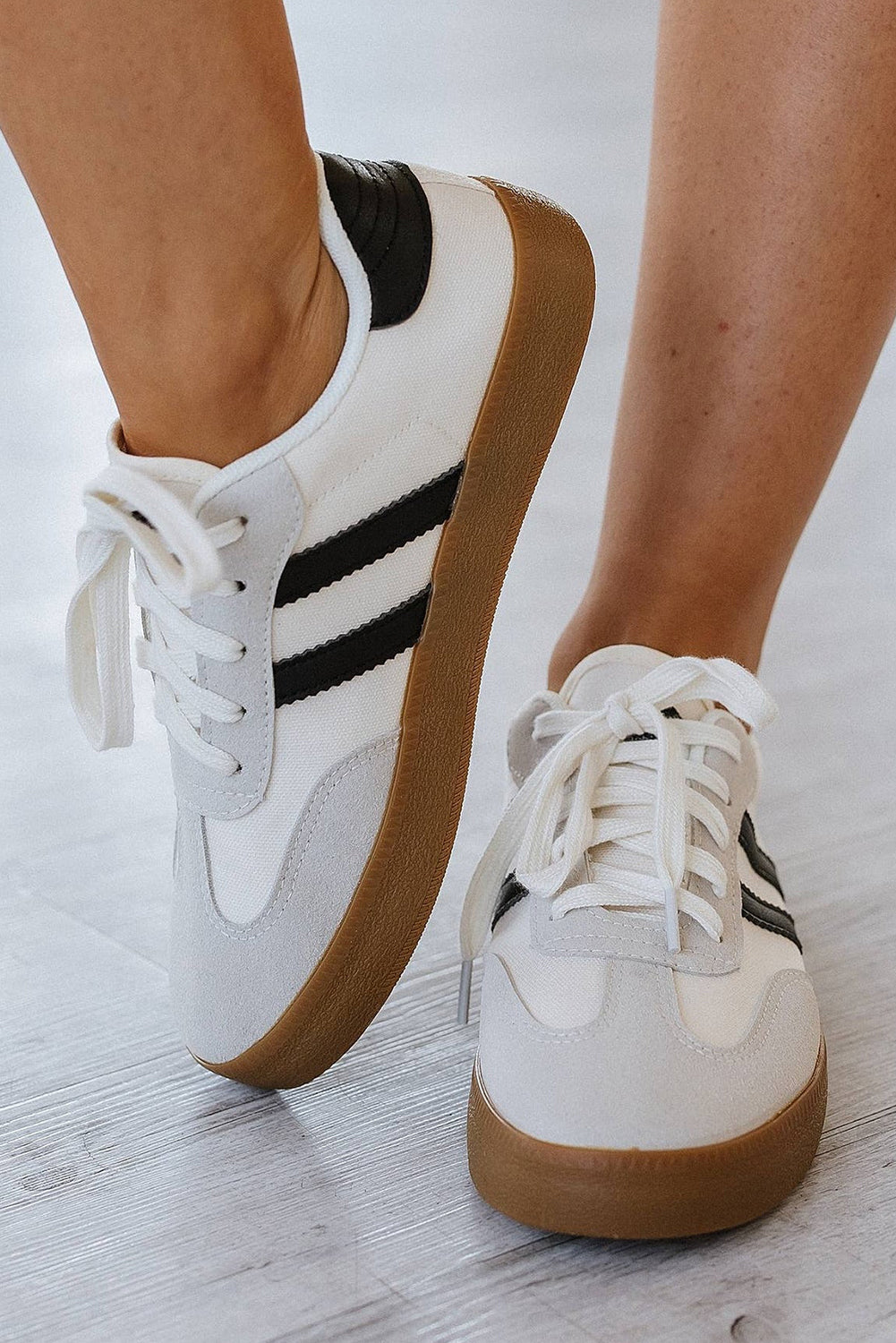 White sneakers with black stripes and brown soles worn by a person on a light wooden floor.