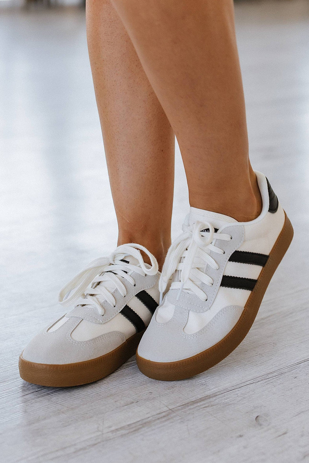 White sneakers with black stripes and brown soles worn by a person on a light wooden floor.