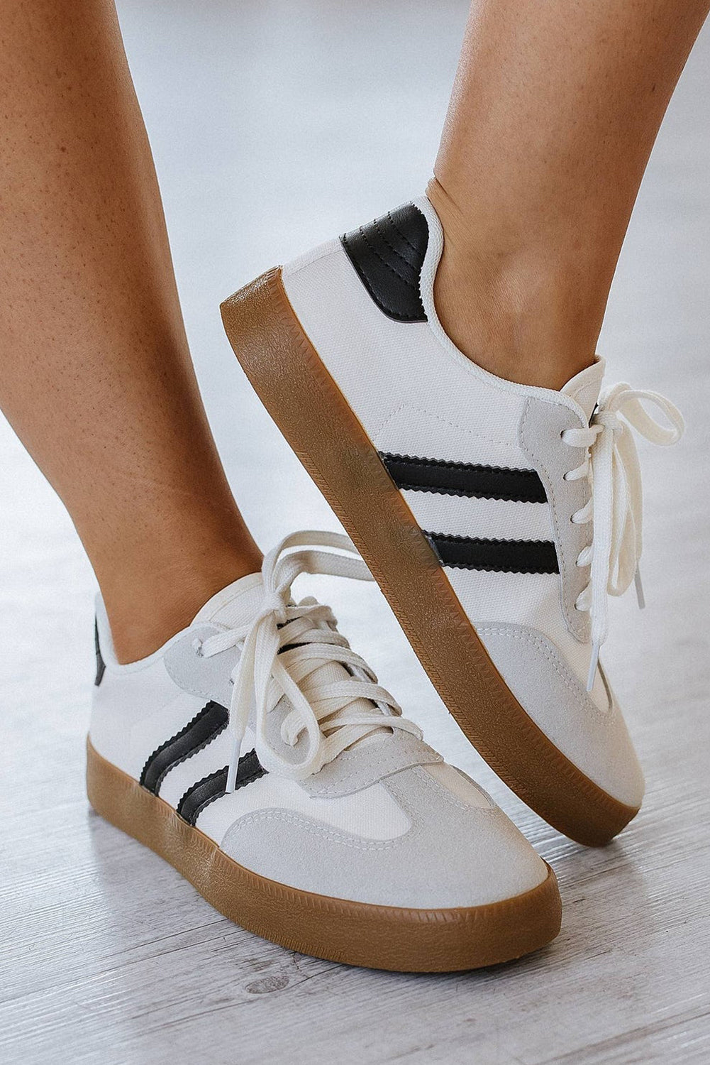 White sneakers with black stripes and brown soles worn by a person on a light wooden floor.