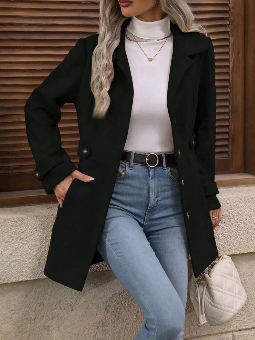 Person wearing a black coat, white turtleneck, and blue jeans against a wooden background
