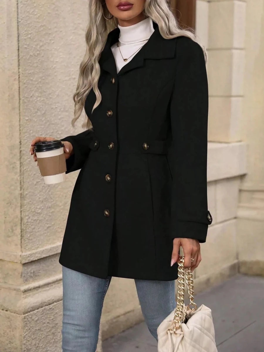 Person wearing a black coat holding a coffee cup and a handbag, standing against a neutral wall.