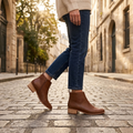 A person wearing brown leather ankle boots with low block heels and side zippers, paired with blue jeans and a cream sweater, walking on a cobblestone street.