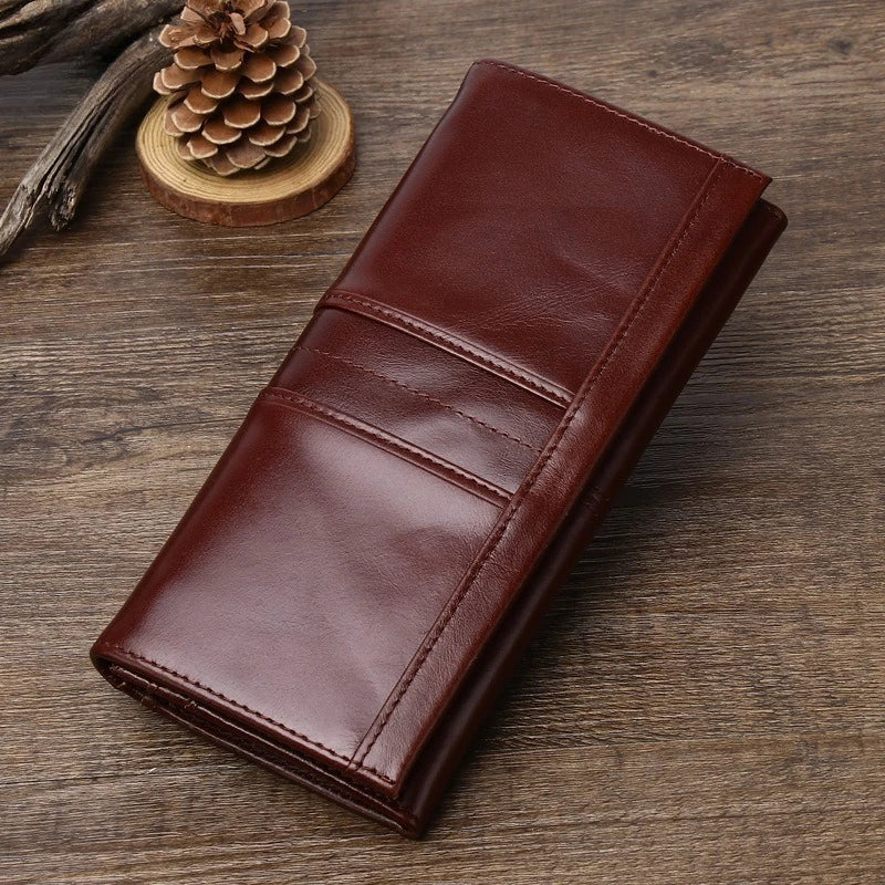 A dark brown leather long wallet or clutch with stitched card slots, displayed diagonally on a rustic wooden surface next to a pinecone.