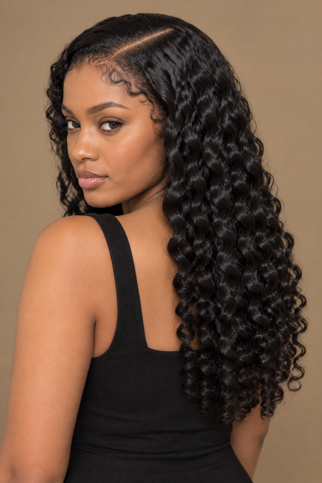 A woman with long, dark, curly hair wearing a black sleeveless top, photographed against a plain beige studio background.