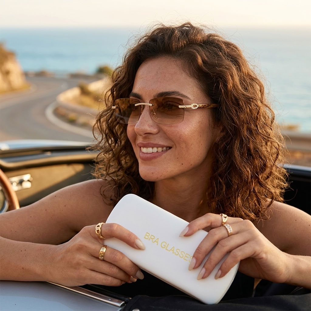A woman with curly brown hair leans in a convertible car by the ocean, wearing gold-framed sunglasses and holding a white rectangular case with gold lettering that reads 'BRA GLASSES'.