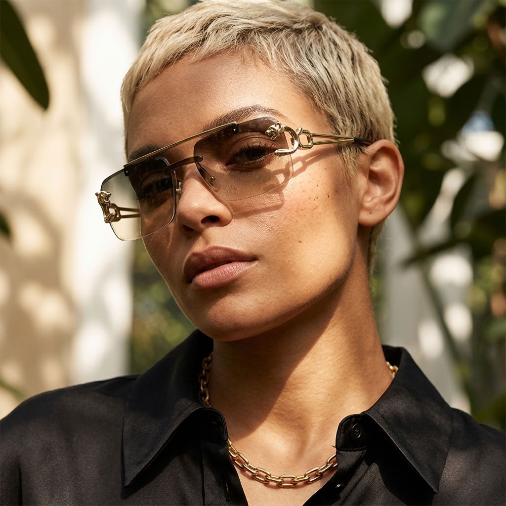 A close-up portrait of a person with short blonde hair wearing stylish square sunglasses with a gold-tone metal frame and clear lenses, paired with a black collared shirt and a gold chain necklace, set against a softly blurred outdoor background with green foliage.
