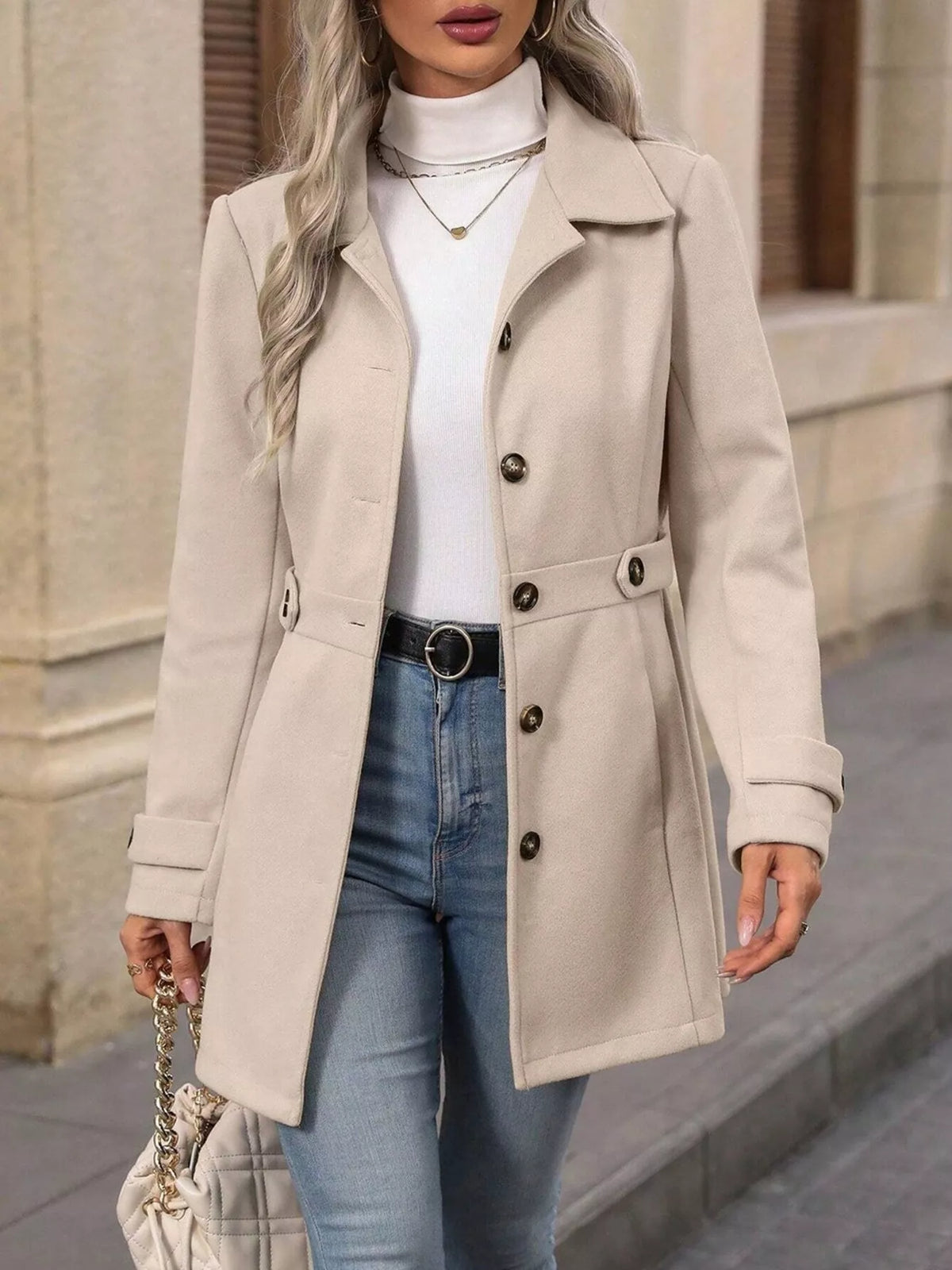 Woman wearing a beige coat, white turtleneck, blue jeans, and holding a handbag on a street.