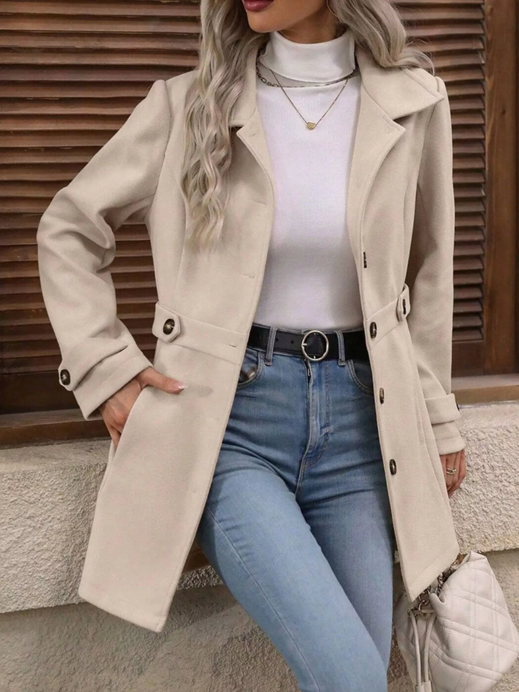 Person wearing a beige coat, white turtleneck, blue jeans, and holding a white bag against a wooden background.