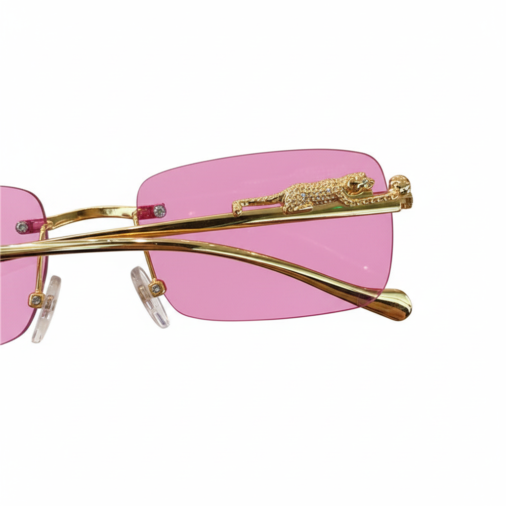 Close-up of rimless sunglasses with pink tinted lenses, a gold metal frame, and a decorative gold crocodile embellishment on the hinge, shown against a white background.