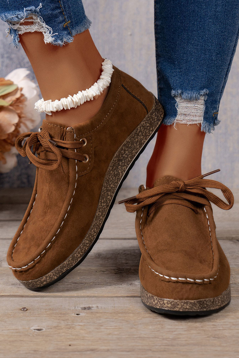 Brown suede shoes with lace-up design worn with ripped jeans on a wooden floor.