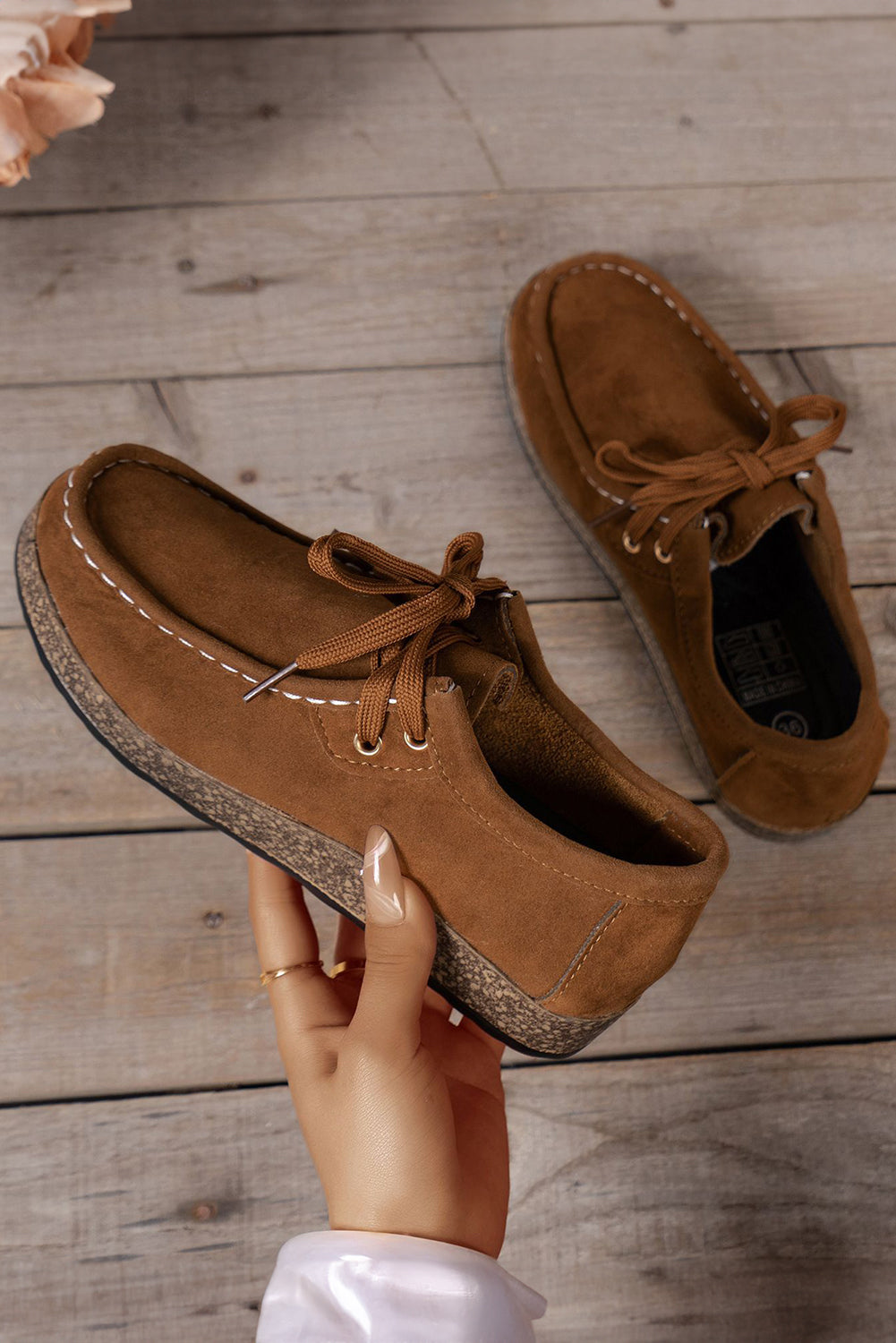 Brown suede shoes on a wooden floor with a hand holding one shoe.