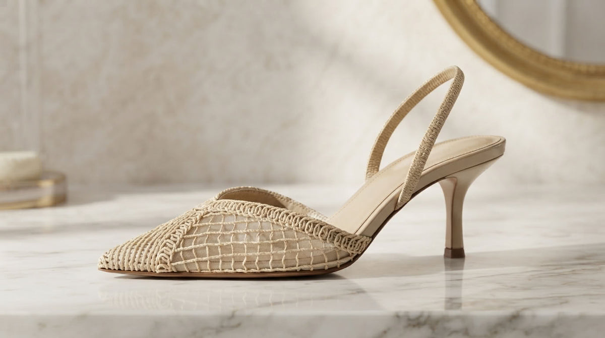 A single beige slingback high-heel shoe with a textured woven upper, displayed on a white marble surface with a warm, softly lit background.