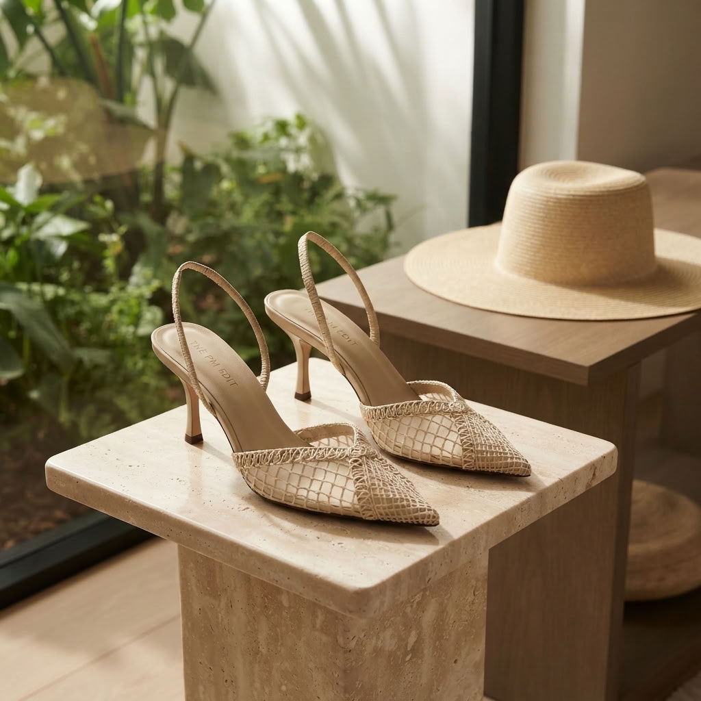 A pair of beige woven slingback kitten heels displayed on a stone pedestal in a sunlit interior, with a straw hat on a wooden table in the background.