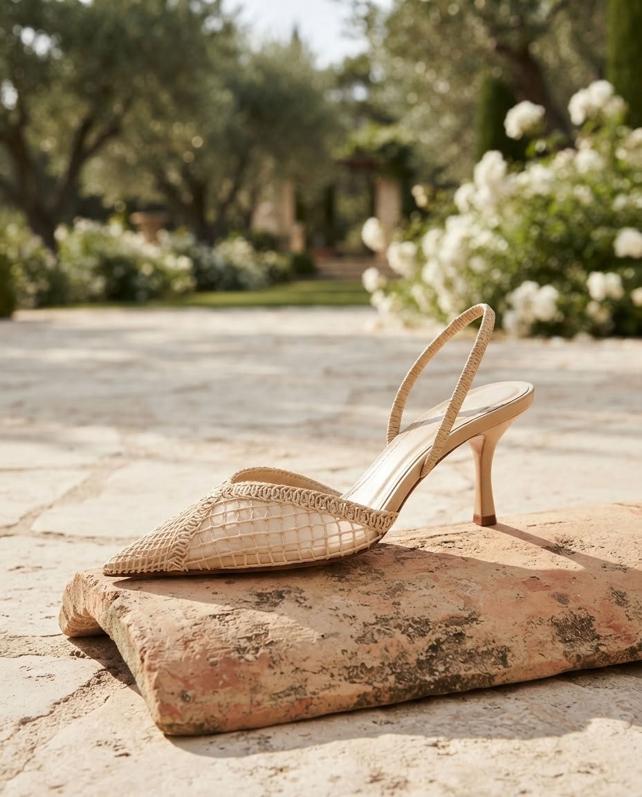 A single pointed-toe high-heel sandal with a woven snakeskin pattern in beige and tan, resting on a rustic stone block in an outdoor garden setting.
