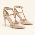 A pair of beige patent leather pointed-toe high heels with gold pyramid stud detailing along the straps, set against a solid cream background.