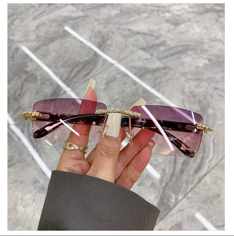 A hand holding a pair of rimless square sunglasses with pink-tinted lenses, gold-toned decorative metal ends, and a tortoiseshell-patterned temple arm, set against a gray marble-patterned background.